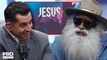 Two men on a PBD Podcast set: a suited host speaking into a microphone and a bearded guest wearing sunglasses and a hat, with a JESUS sign in the background.